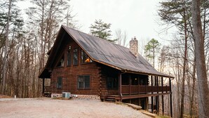 A log cabin with a porch, a stone chimney, and a gravel driveway.