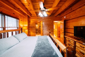 Interior de una cabaña de madera con una cama, un ventilador de techo, un televisor y paredes de madera.