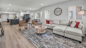 Ein modernes Wohnzimmer mit Sofa, Couchtisch und Sesseln. Es gibt eine Küchenzeile mit Kühlschrank und einem Esstisch.
