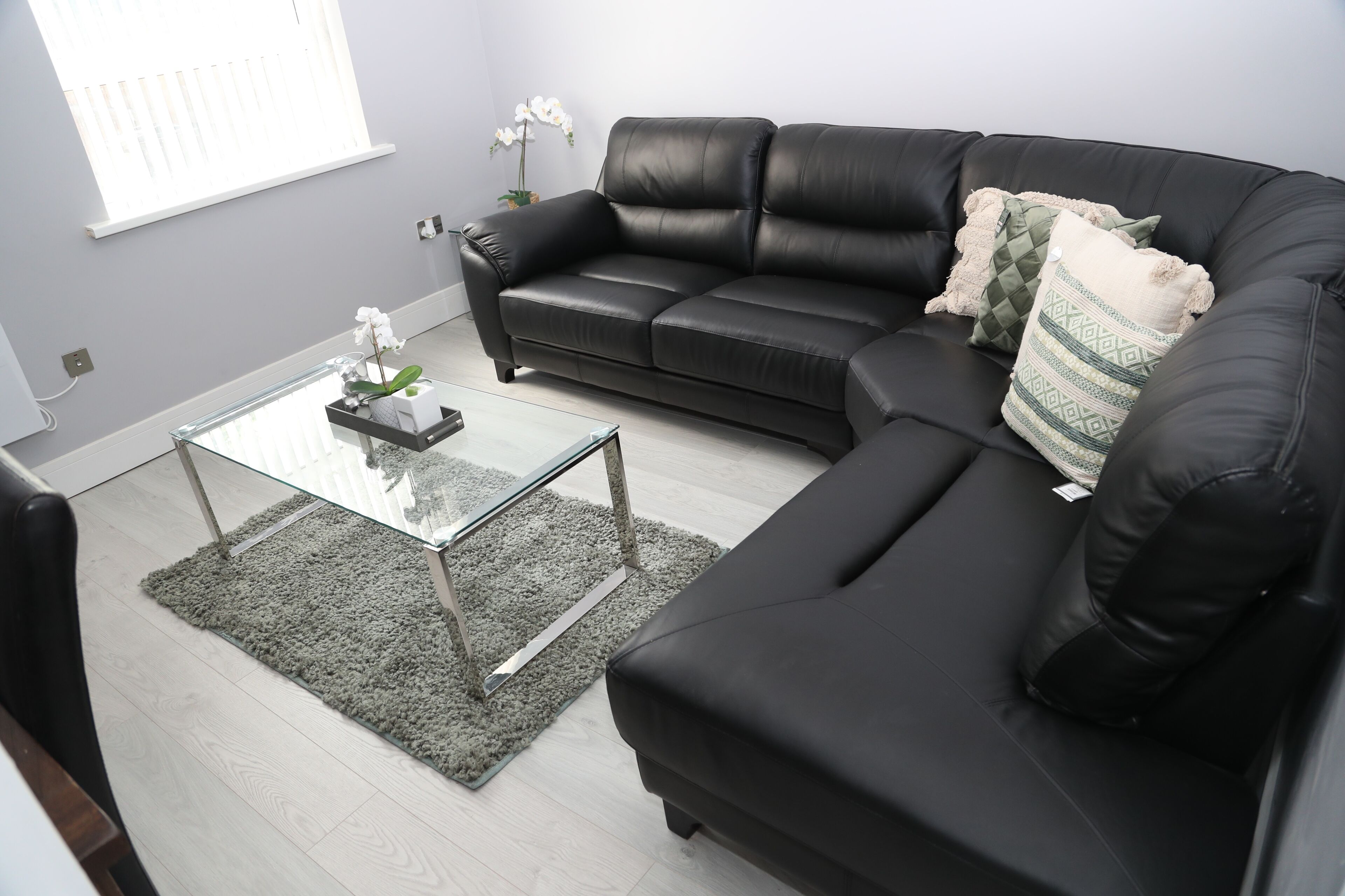 A modern living room with a black leather sofa, a glass coffee table, and a grey rug.