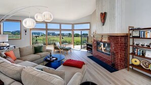 A spacious living room with a fireplace, large windows, and a view of the outdoors. - Pacific Flyaway, by Vinifera Homes (Bodega Bay)