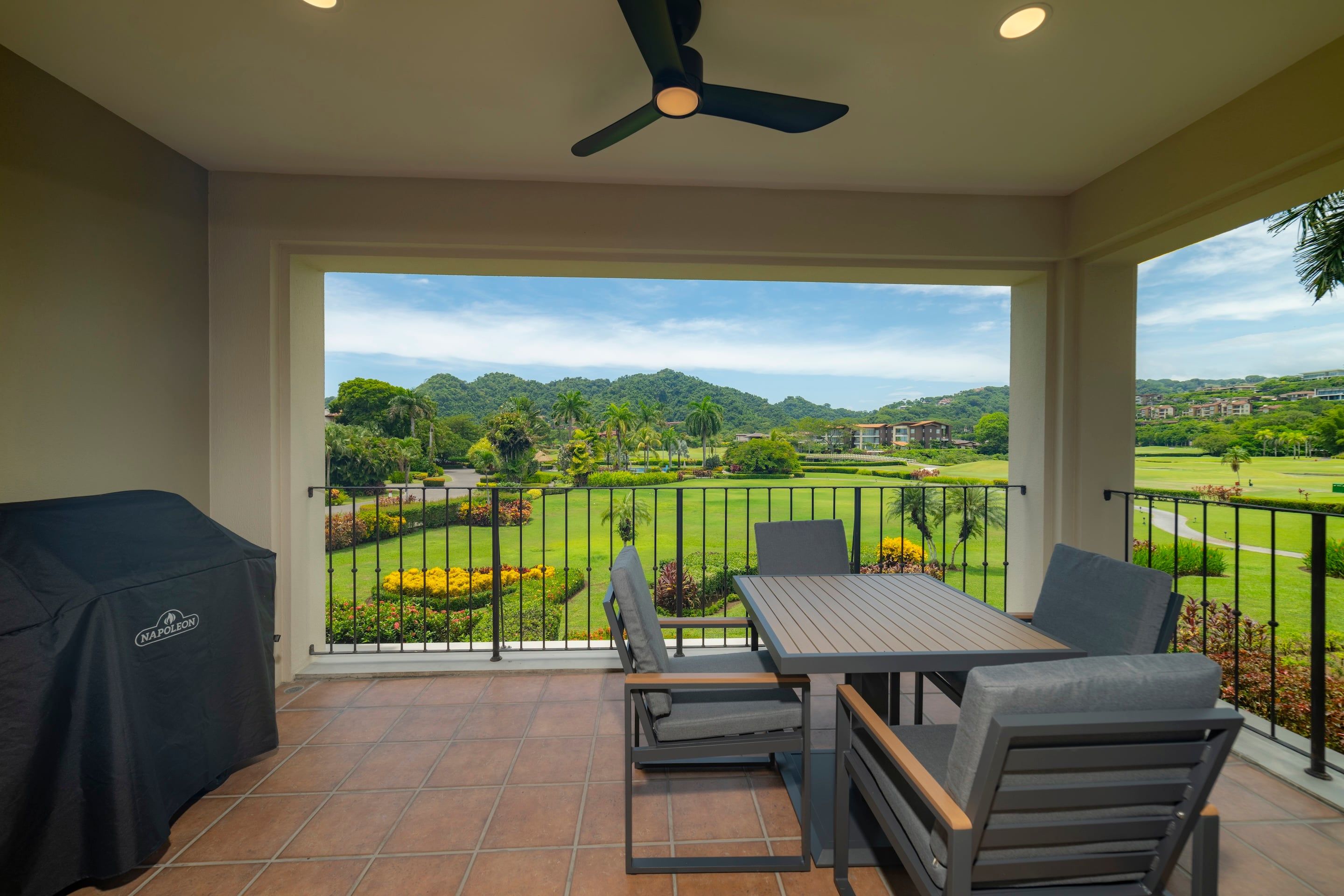 A balcony with a table and chairs, a ceiling fan, and a view of a landscaped area with buildings in the distance.