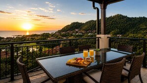 En balkon med et glasbord, fletstole og havudsigt i solnedgangen.