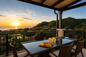 Ein Balkon mit Glastisch, Korbstühlen und Blick auf den Sonnenuntergang über dem Meer.