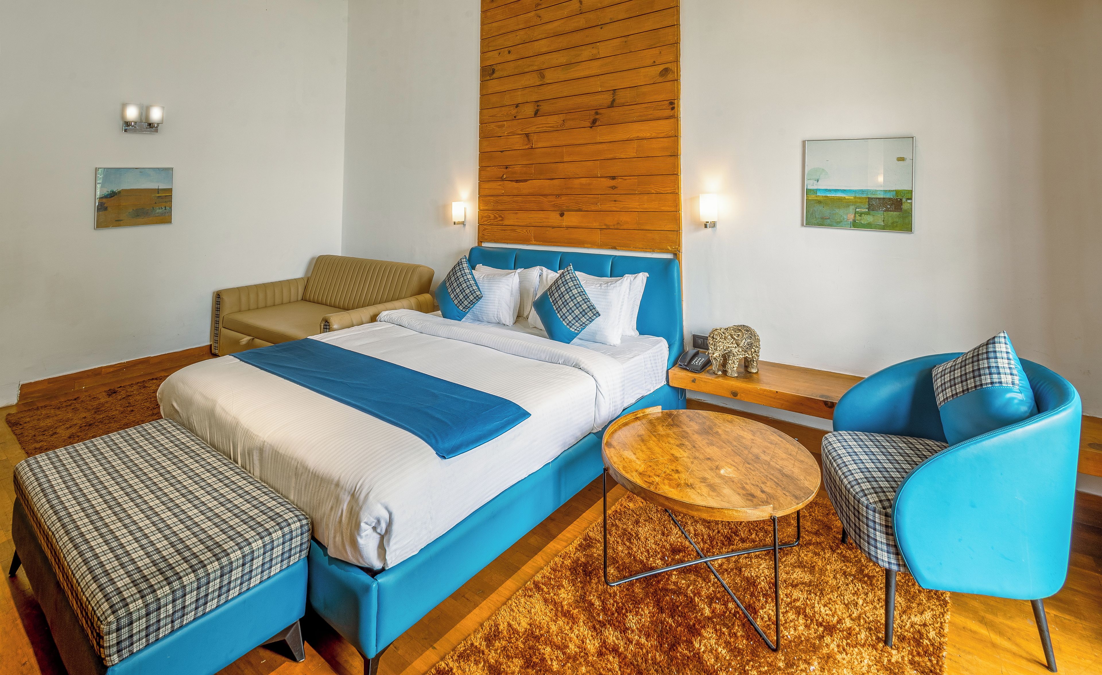 A hotel room with a bed, a sofa, a chair, a small table, and artwork on the walls.