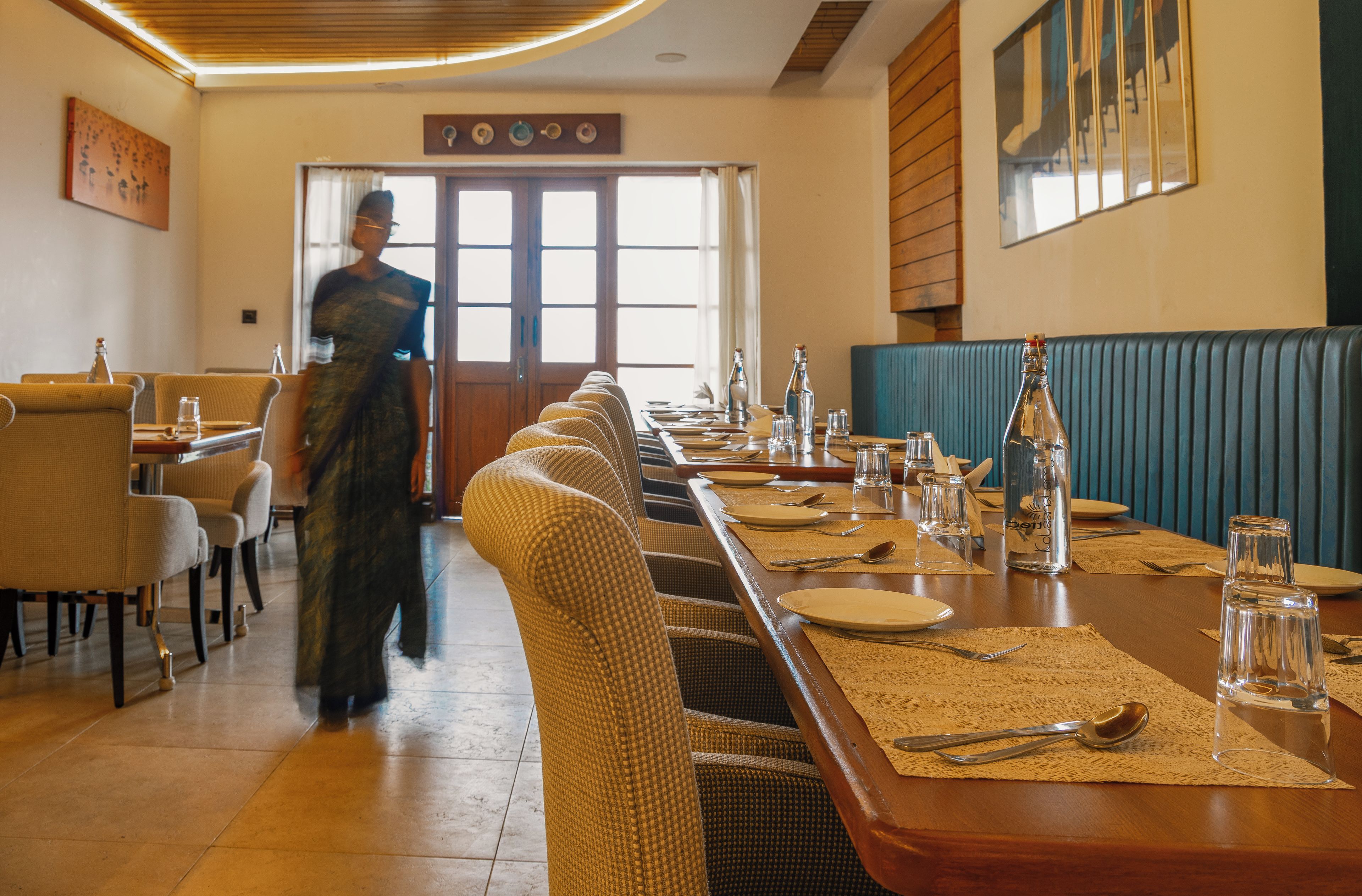 A restaurant with a long dining table set for a meal, chairs, and a person walking through the space.