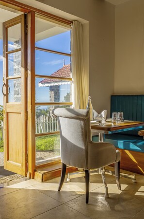 A room with a view of a building with a red roof, a wicker chair, a dining table with glasses, and a pool.