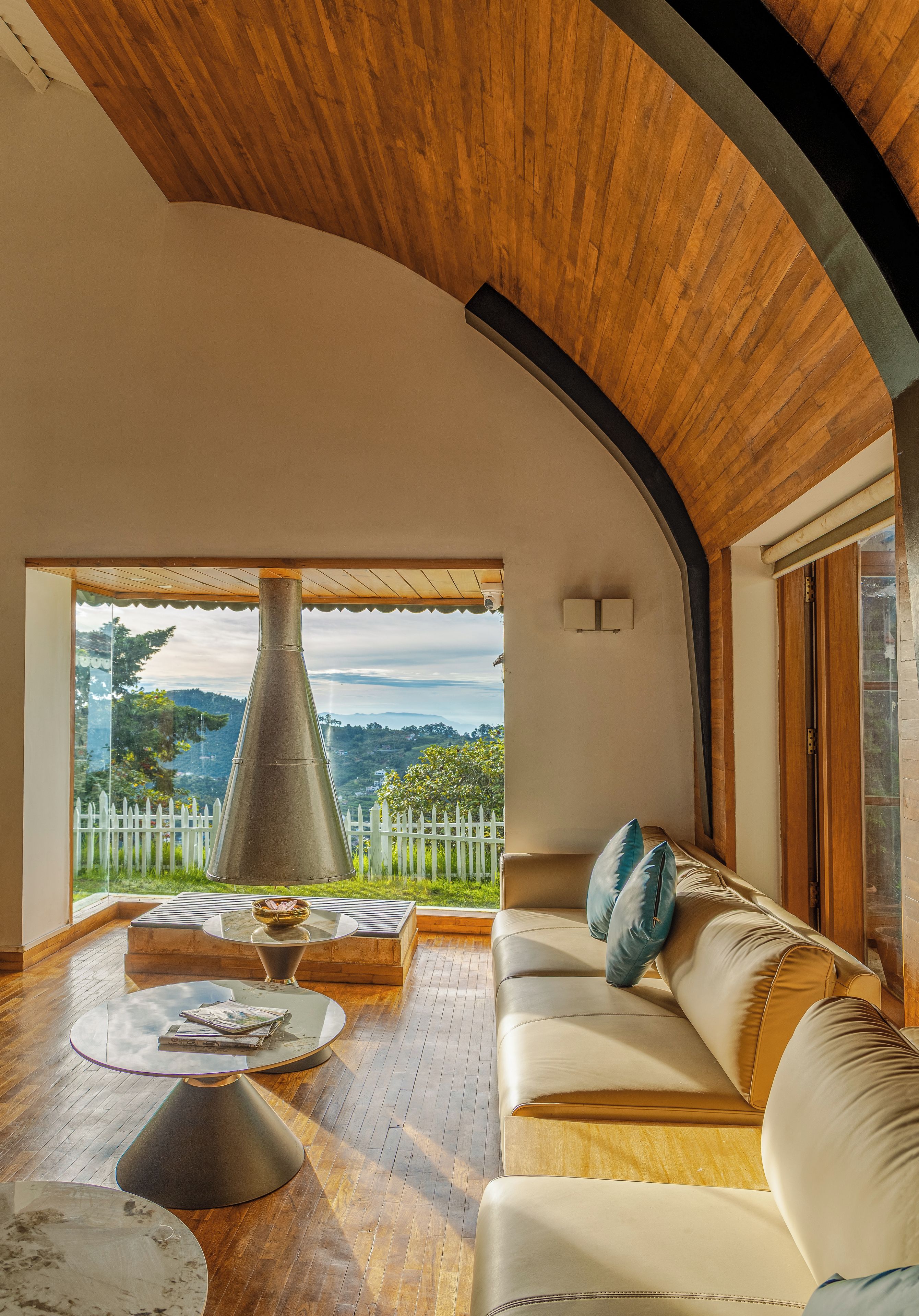 A spacious living area with a wooden ceiling, a sofa, a coffee table, and a view of a white picket fence and greenery.