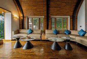 A spacious living area with a high ceiling, wooden flooring, and large windows offering a view of greenery. - The Mainland Resorts Kodaikanal (Kodaikanal)