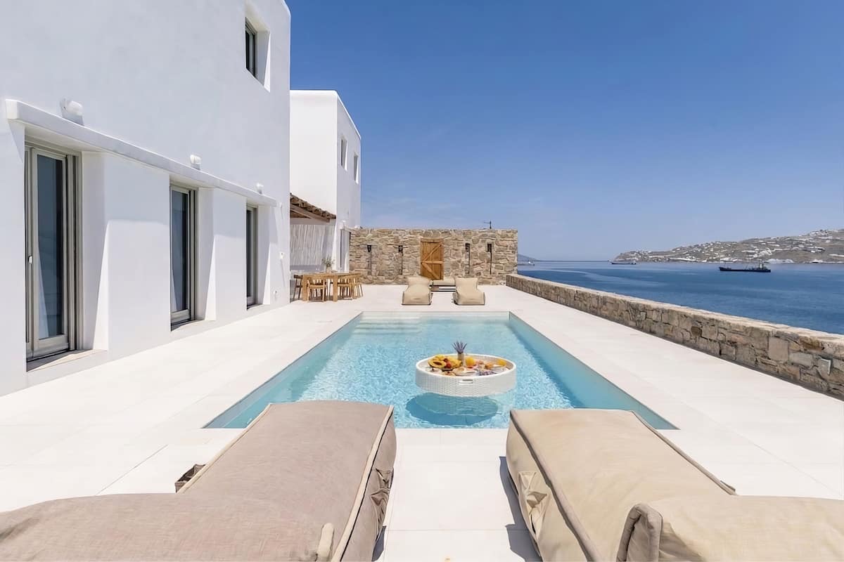 A modern pool area with lounge chairs, a stone wall, and a view of the sea.