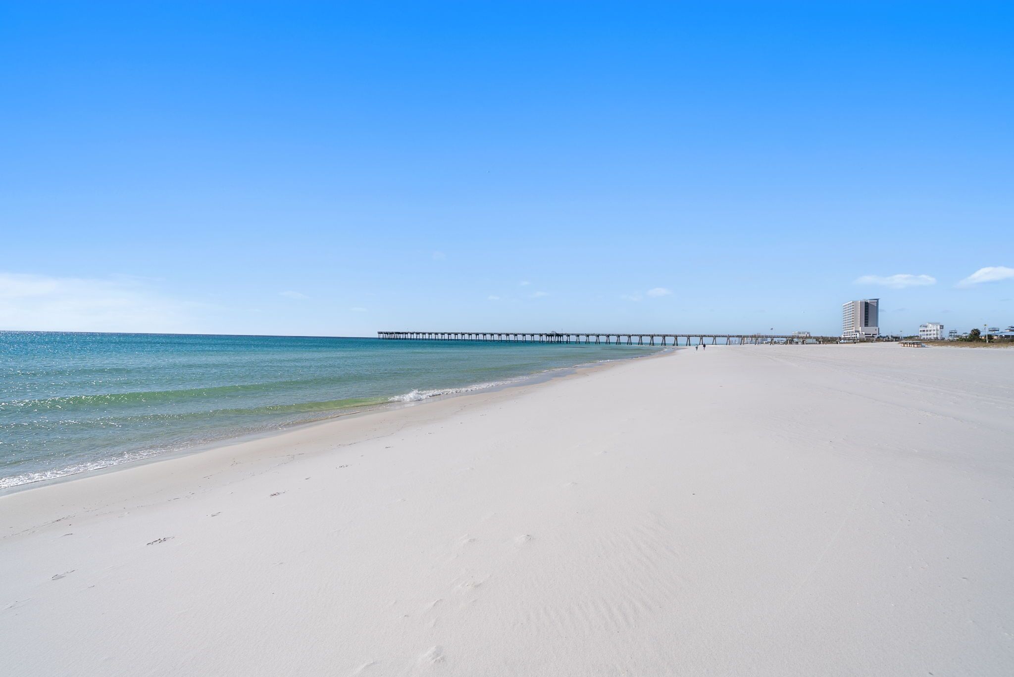 A beach with a pier extending into the ocean, a clear sky, and buildings in the distance.
