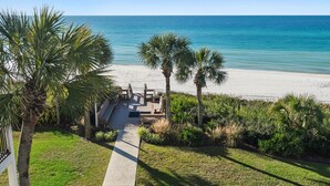 A beach view with a pathway leading to a wooden deck, palm trees, and a clear blue sky.