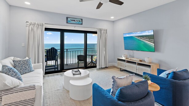 A modern living room with a balcony, white sofas, blue armchairs, a flat-screen TV, and a ceiling fan.