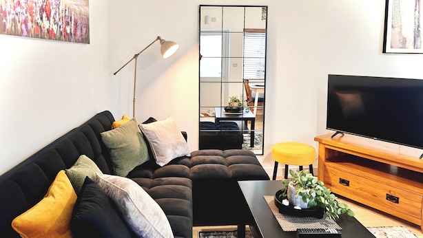 A modern living room with a black sofa, a wooden TV stand, a floor lamp, and a large mirror. - Charming Apartment Nottingham FREE onsite parking (Nottingham)