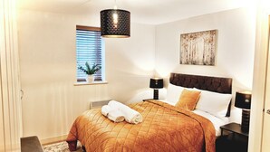 A neatly made bed with a brown quilt, a bedside lamp, a small table, and a window with blinds. - Charming Apartment Nottingham FREE onsite parking (Nottingham)