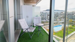 Un balcón con techo de madera, sillas blancas, una mesita y vistas a una piscina y edificios.