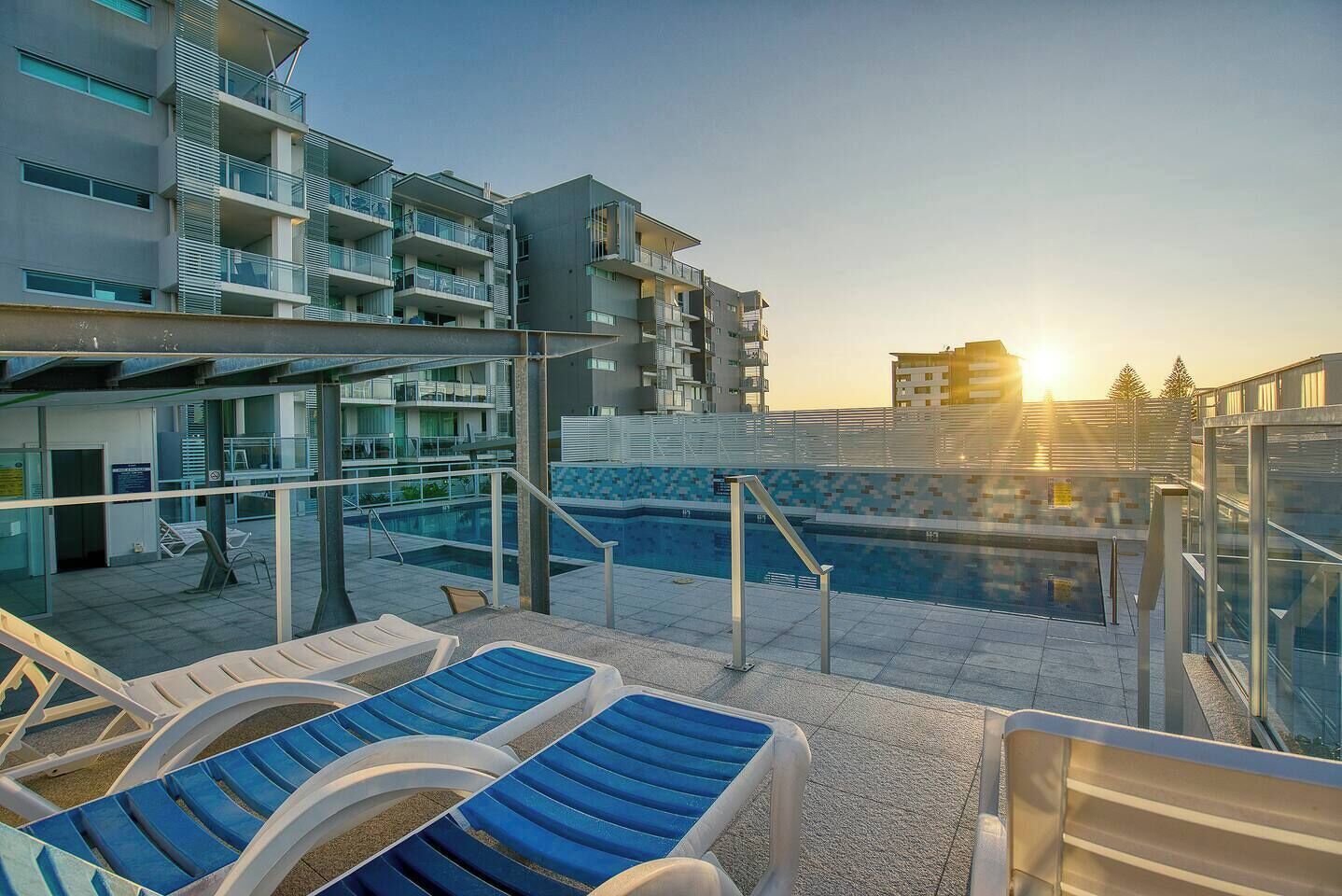 Uma área de piscina no terraço com espreguiçadeiras, piscina e edifícios modernos ao fundo.
