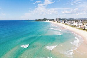 A coastal beach with clear turquoise waters, a sandy shore, and a row of buildings along the coastline. - Pure Palm Beach Bliss at The Pavilions on Fifth (Palm Beach)