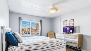 Ein Schlafzimmer mit Bett, Schreibtisch, Sessel, Deckenventilator und Fernseher an der Wand.