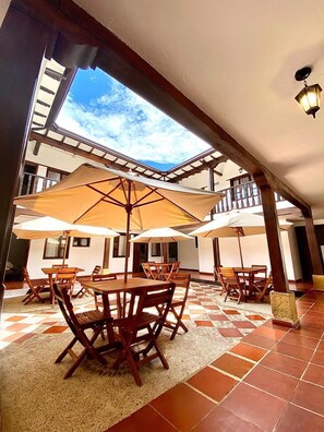 Un patio exterior con mesas y sillas de madera, sombrillas grandes y vistas al cielo a través de un tragaluz.