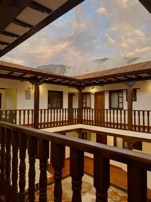 A courtyard with a wooden balcony railing, a tiled floor, and a view of mountains under a cloudy sky.
