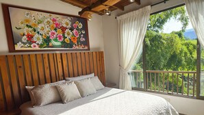 Ein Schlafzimmer mit einem hölzernen Kopfteil, einem großen Bett mit einer Steppdecke, einem Blumenbild an der Wand und einem Balkon mit Blick ins Grüne.