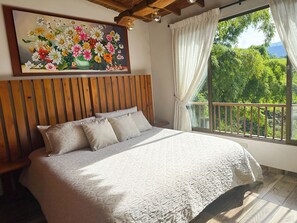 Una camera da letto con testiera in legno, un letto grande con copriletto e un quadro di fiori sulla parete, e un balcone con vista sul verde.