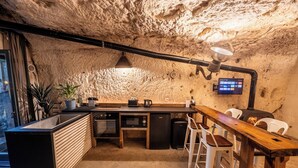 Una cocina con pared de piedra, una mesa de madera, taburetes de bar, una cafetera y un refrigerador pequeño.
