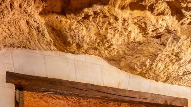 Un dormitorio con techo de piedra rústica, cama con ropa de cama roja, ventana redonda y puerta de madera.