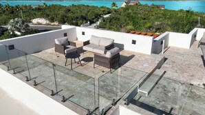 Terraço no telhado com guarda-corpo de vidro, mobiliário para área externa e vista para um mar azul-turquesa e paisagem tropical.