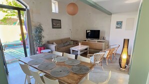 Uma área de refeições com mesa de madeira e cadeiras brancas, um sofá, uma televisão e um vaso de planta.