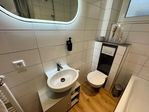 Une salle de bain moderne avec un lavabo, des toilettes et un meuble-lavabo blancs. Un miroir est placé au-dessus du lavabo, ainsi qu’un distributeur de savon liquide et une petite poubelle.
