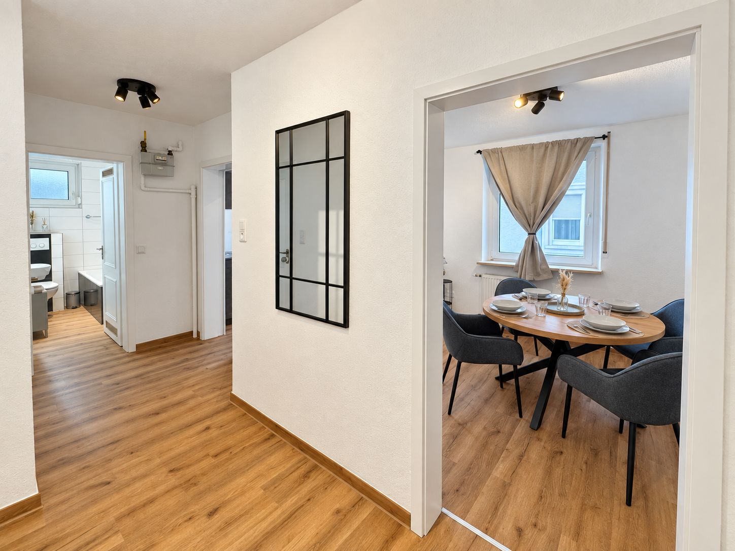 A modern interior with a wooden floor, a dining area with a round table and chairs, and a kitchen visible in the background.