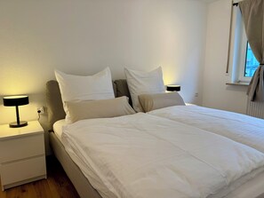 Un lit bien fait, avec du linge de lit blanc, une lampe de chevet et une table de chevet dotée d’un tiroir.