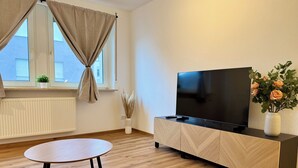 Un salon moderne avec un plancher en bois, une table ronde, un téléviseur posé sur un meuble, une fenêtre avec des rideaux et un vase rempli de fleurs.