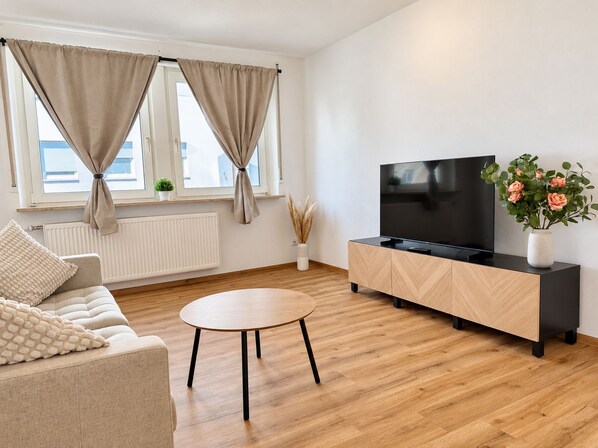 Un salon moderne avec un canapé, une table basse ronde, un téléviseur posé sur un meuble et une fenêtre avec des rideaux.
