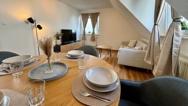 Un moderno comedor con una mesa de madera dispuesta para cuatro, un televisor al fondo y una zona de estar acogedora con un sofá y cortinas.