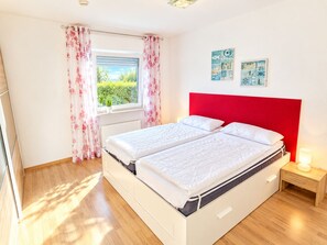 A double bed with a red headboard, a bedside table with a lamp, and a window with curtains. - Ground-floor vacation apartment (Altenkirchen)