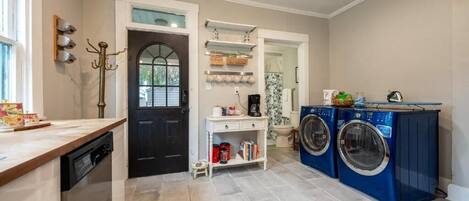 A laundry room with a dishwasher, two washing machines, a coffee maker, and a small shelf with books and supplies.