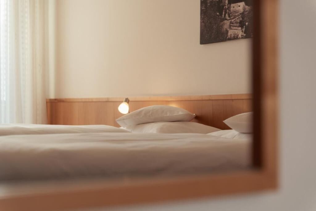 A neatly made bed with white linens and pillows, a wooden headboard, and a wall-mounted light fixture.