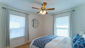 Ein Schlafzimmer mit einem Bett, einem Deckenventilator, einem runden Spiegel und großen Fenstern mit Vorhängen.