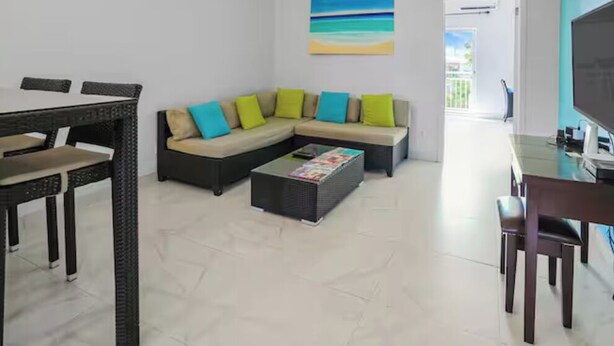 Ein Wohnzimmer mit Ecksofa, Couchtisch, Esstisch mit Stühlen und Fernseher.