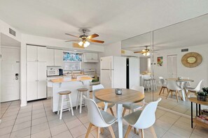 Une cuisine moderne avec des meubles blancs, un îlot central doté de tabourets et un coin repas avec des chaises blanches et une table en bois.
