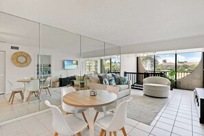 A modern living room with a round wooden table, white chairs, a sofa with cushions, a TV, and a large window offering a view of the outdoors. - Prime Marco Island Condo Close to the Beach (Marco Island)