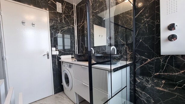 A modern bathroom with a black marble backsplash, a glass shower enclosure, a built-in washing machine, and a white vanity with a mirror. - 3-bedroom apartment between Paris and Disneyland - 82m3 - Close to transport links (Neuilly-sur-Marne)
