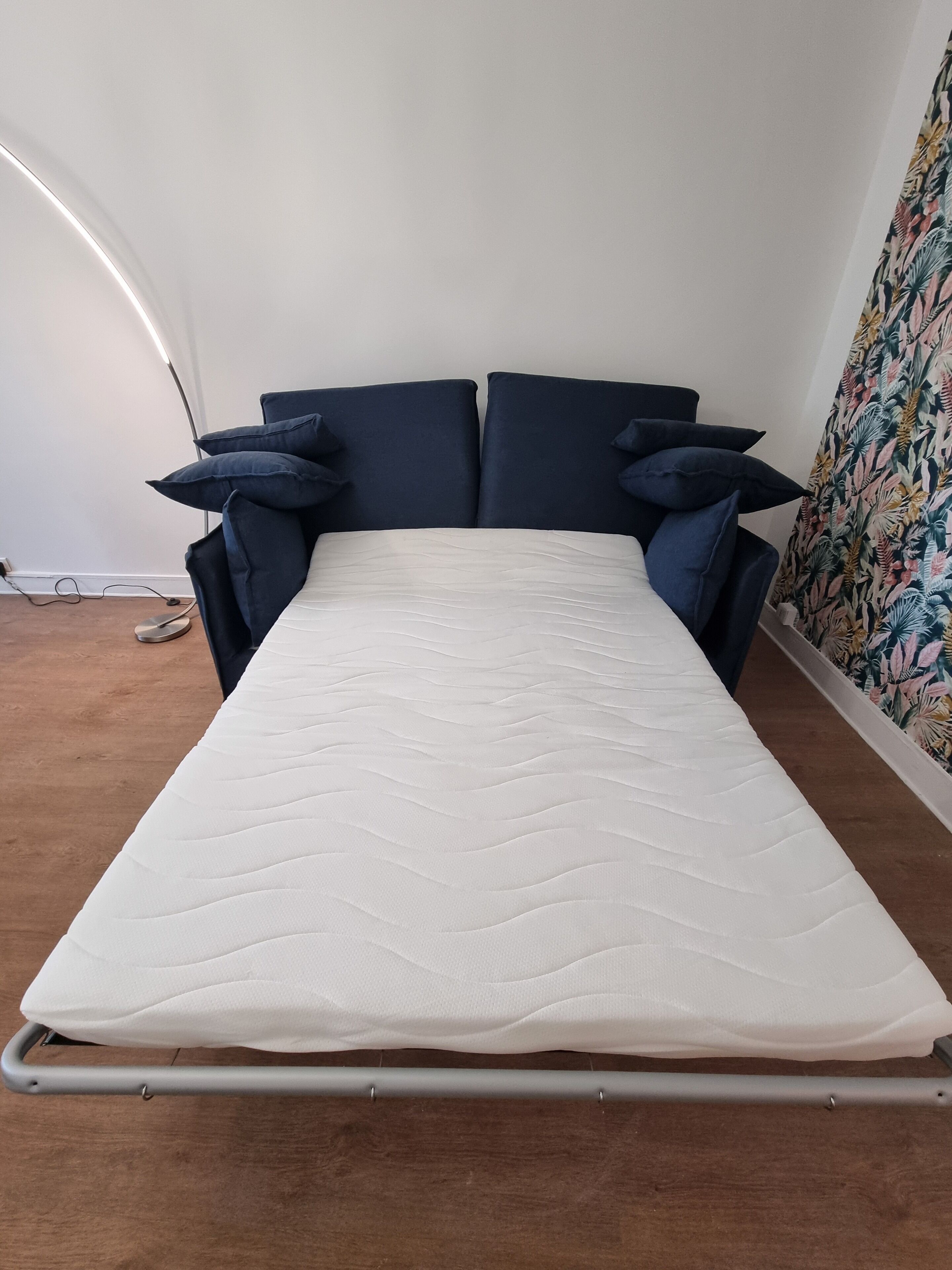 Uma cama de solteiro com colchão branco, cabeceira e peseira em azul escuro, posicionada em frente a uma parede lisa com uma cortina estampada à direita.