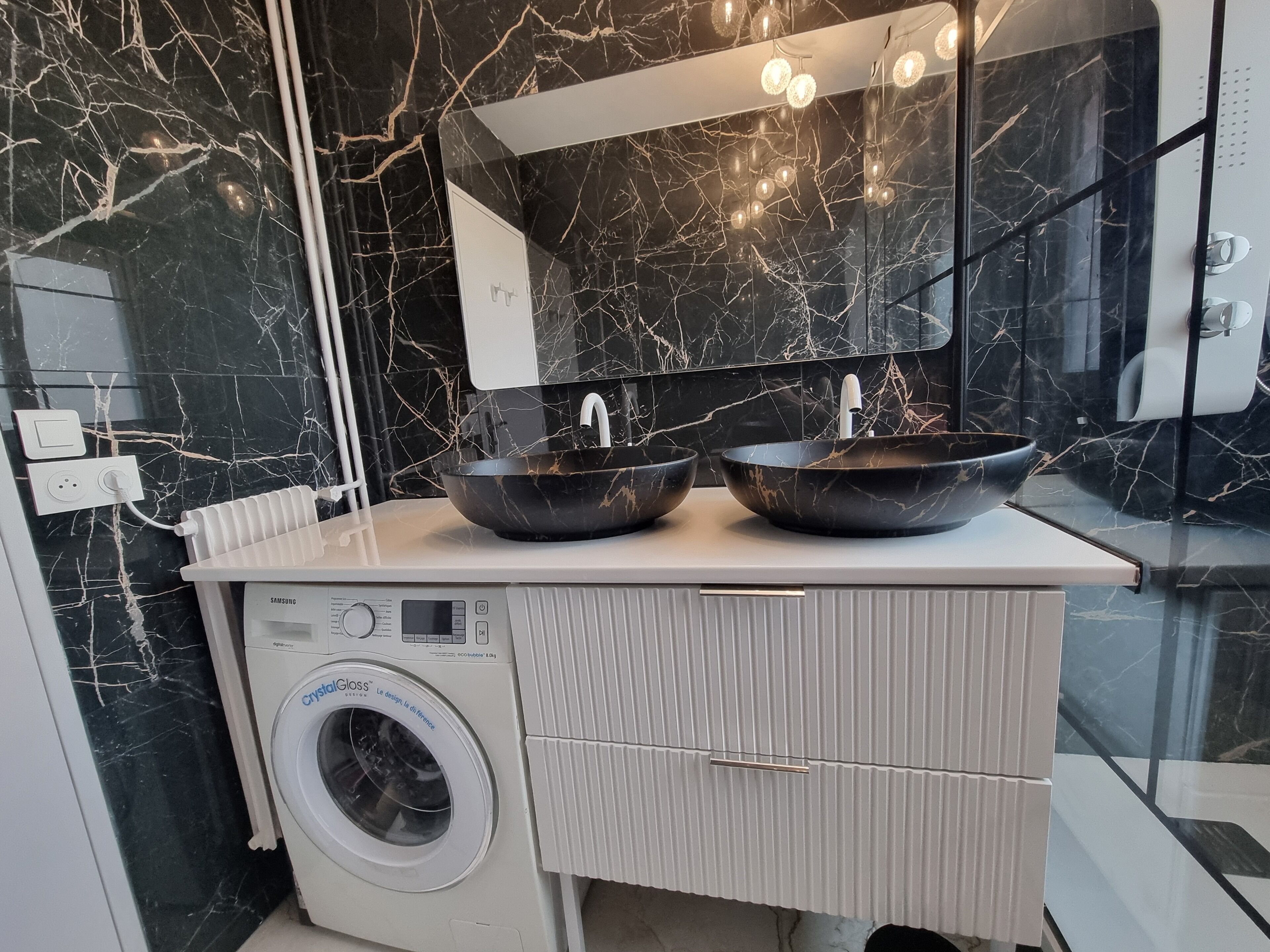 Un baño moderno con una encimera de mármol negro, dos lavamanos negros tipo recipiente, una lavadora blanca y un espejo grande sobre los lavamanos.