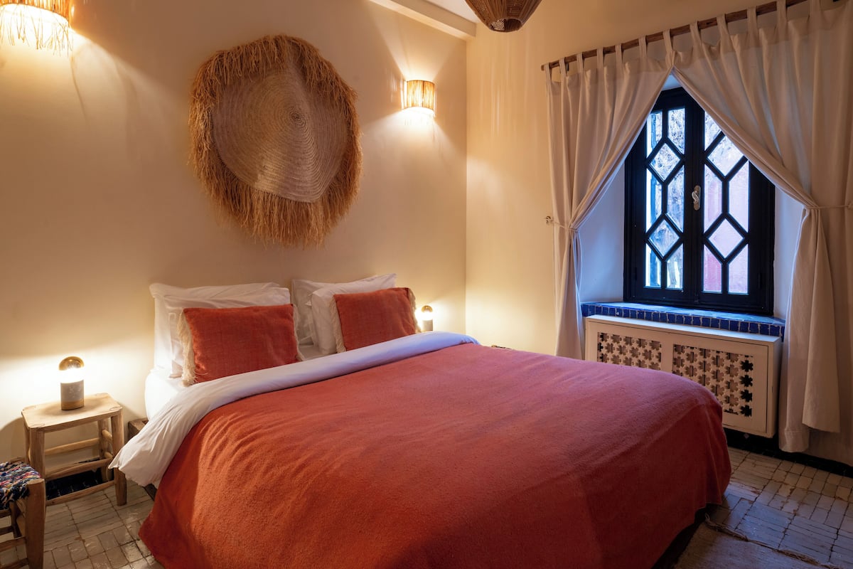 A bed with a red blanket, a wall-mounted hat, and a window with a geometric pattern.