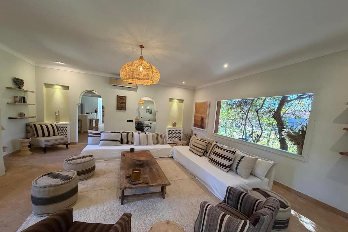 A spacious living room with a large sectional sofa, a wooden coffee table, and striped armchairs. There is a large window with a view of trees.