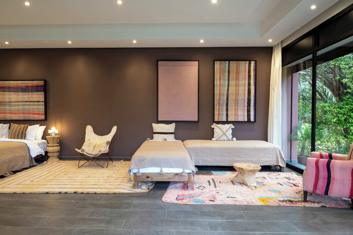 A modern bedroom with a large bed, a smaller bed, a chair, a side table, and a large window with a view of greenery.